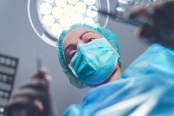 From below woman surgeon in medical uniform using professional tools while standing under bright light in operating theater. High quality photo. From below woman surgeon in medical uniform using professional tools while standing under bright light in operating theater