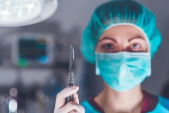 Adult woman in medical mask and hat and with scalpel looking at camera before performing surgery in hospital. High quality photo. Adult woman in medical mask and hat and with scalpel looking at camera before performing surgery in hospital