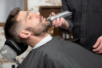 Young hipster Caucasian man during beard grooming in modern barber shop. Men’s hair styling. Handsome man getting new hairstyle with electric trimmer. High quality photography. Young hipster Caucasian man during beard grooming in modern barber shop. Men’s hair styling. Handsome man getting new hairstyle with electric trimmer. 