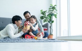 Asian young LGBTQ gay family giving gift to little adopted daughter on birthday or special day in living room at home. Caucasian kid girl opening present box with smiling and surprising face