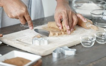 Selective focus on senior man hands taking small trowel to remove gingerbread cookie dough from board after pressed mold on dough at kitchen with flour, cinnamon powder on wooden table at home