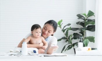Cute Caucasian little naked toddler baby girl sitting on towel after bathing and wipe body dry while young Asian mother apply talcum powder on her kid skin at home. Hygiene care for children concept