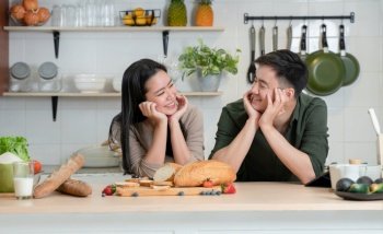 Portrait of young Asian romantic couple smiling, placing hands on chin, looking at each other in the kitchen, happy beautiful wife and handsome husband cooking healthy breakfast food together at home