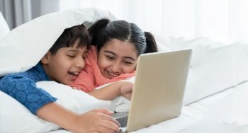 Two happy Indian brother and sister in traditional clothing lying on bed under blanket smiling, using laptop, having fun together at home. Education, Siblings relationship concept