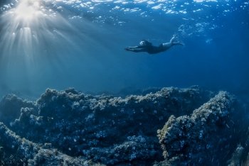 Girl swimming underwater in a rocky bottom with sunbeams coming from the left. Freediving in the Mediterranean sea