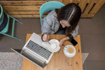 From above of female freelancer sitting at wooden table and eating sweet dessert and drinking coffee while working on laptop. Woman eating dessert and drinking coffee during online work