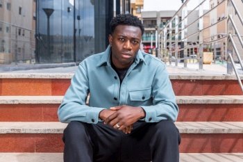 Calm trendy young African American male millennial with curly dark hair in stylish outfit sitting on stairs near entrance of contemporary building and looking at camera. Confident ethnic guy sitting on stairs on street