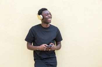 Happy young African American male millennial with dark curly hair in casual clothes smiling and looking away while using smartphone and listening to music in modern headphones. Delighted young black man listening to song in headphones and surfing smartphone