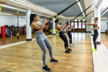 Female coach explaining exercise to sportswomen hanging on TRX ropes during suspension workout in spacious light gym. Trainer showing exercise to athletes during suspension training