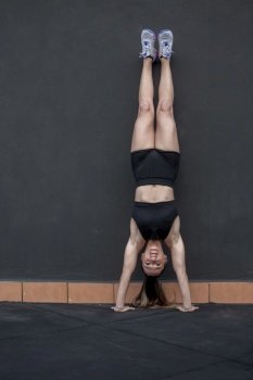 Full body strong sportswoman in sportswear doing handstand near black wall during functional fitness training in gym. Female athlete doing handstand exercise