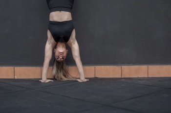 Ground level of muscular sportswoman in activewear doing handstand exercise near black wall during fitness workout in gym. Female athlete doing handstand during training