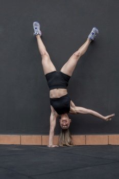 Full body female athlete in sportswear doing one arm handstand near wall during intense fitness training in gym. Sportswoman doing one arm handstand
