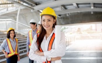 Portrait of Asian beautiful female engineer wearing safety helmet and uniform while standing in construction site with blur background of her colleagues