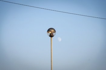Light pole at sunset in front of a blue sky with the moon, the pole is hit by the last rays of sunlight of the day, there is also an electric cable.
