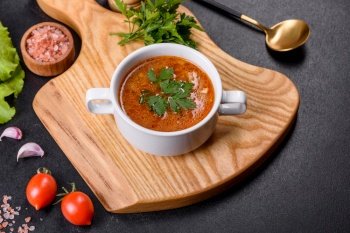 Fresh delicious tomato soup with spices and parsley in a white bowl on a wooden board against a dark concrete background. Fresh delicious tomato soup with spices and parsley in a white bowl on a wooden board