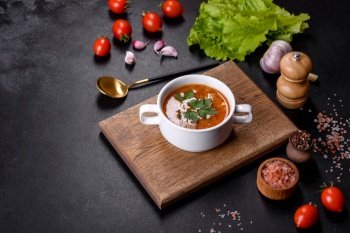 Fresh delicious tomato soup with spices and parsley in a white bowl on a wooden board against a dark concrete background. Fresh delicious tomato soup with spices and parsley in a white bowl on a wooden board