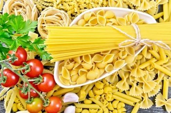 Pasta seashells and spaghetti in bowl, pipe rigate, tagliatelle, fusilli, penne, farfalle, conquilla, trenette, rotini, pappardelle, tomatoes, garlic and parsley on wooden board background from above
