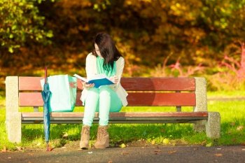 Woman enjoying beautiful autumn nature weather, reading book sitting on bench in park during sunny autumnal day,. Woman reading book sitting on bench in park