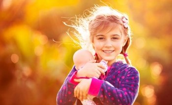 Portrait of a cute little girl holding in hands her favorite precious doll, playing outside, over natural orange background, pretty child playing mother’s daughters in warm autumn day