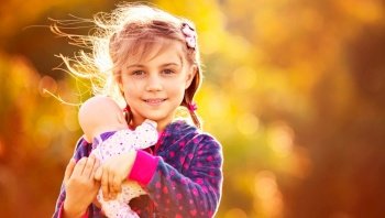 Portrait of a sweet baby girl with doll over orange autumnal foliage background, happy child playing mother’s daughters outdoors in fall sunny day