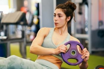 Sporty young female, wearing sportswear, on yoga mat doing situps with plate weights in gym. Fitness woman doing abs crunches.. Young sportswoman on yoga mat doing situps in gym.