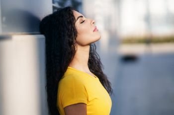 Young Arab woman with her eyes closed and her head leaning against the wall in the street. Curly hair female.. Arab woman with eyes closed in urban background