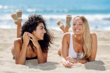 Two young women with beautiful bodies in swimwear on a tropical beach. Funny caucasian and arabic females wearing black and white swimsuits lying on the sand on the beach. Two young women with beautiful bodies in swimsuit on a tropical beach