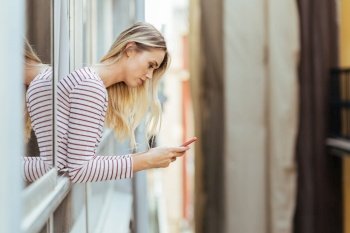 Attractive woman leaning out of her house window using a smart phone.. Attractive woman leaning out of her house window using a smartphone.
