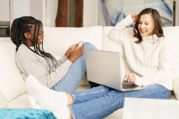 Two female student friends sitting on the couch at home using a laptop. Multiethnic women.. Two female student friends sitting on the couch at home using a laptop.