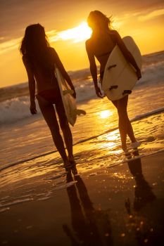 Rear view of two beautiful sexy young women surfer girls in bikinis with white surfboards on a beach at sunset or sunrise     Two Women Bikini Surfers With Surfboard Sunset or Sunrise Beach