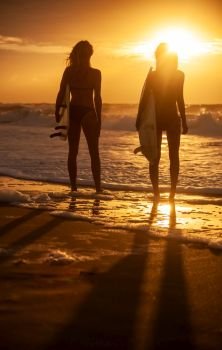 Rear view of two beautiful sexy young women surfer girls in bikinis with white surfboards on a beach at sunset or sunrise 