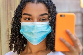 Mixed race African American teenager teen girl young woman wearing a face mask and using a mobile cell phone for social media or selfies during the Coronavirus COVID-19 pandemic