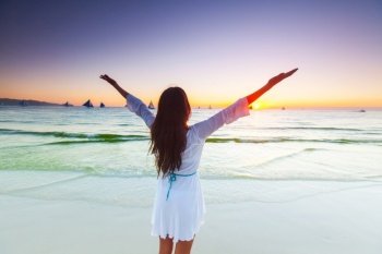 Woman standing on tropical sea beach with raised arms up and looking into a wide sea view with sail boats. Woman raising arms to sunrise