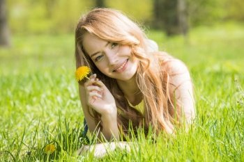 Happy smiling young woman laying on the green grass holding yellow dandelion flower. Woman on the green grass