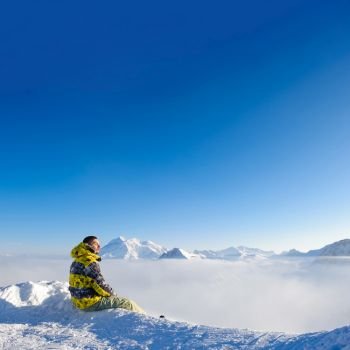 Alpine winter mountain landscape with man sitting above low clouds. French Alps covered with snow in sunny day. Val-d’Isere, France