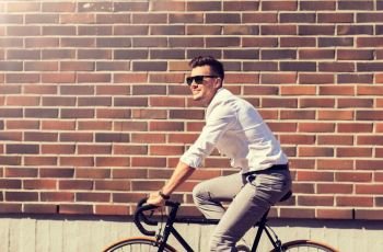 lifestyle, transport and people concept - young man in sunglasses riding bicycle on city street over brickwall. young man riding bicycle on city street