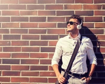 style, music, technology and people concept - happy smiling young man in headphones and sunglasses with bag over brickwall. young man in headphones with bag over brickwall