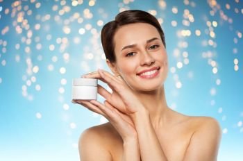 beauty, cosmetics and skincare concept - happy young woman holding jar of cream over holidays lights on blue background. happy young woman holding jar of cream