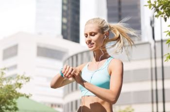sport, technology and people concept - smiling young woman with fitness tracker and earphones exercising over city street on background. happy woman with fitness tracker and earphones