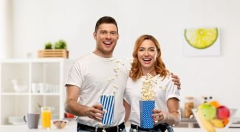food, entertainment and people concept - portrait of happy couple in white t-shirts eating popcorn over home kitchen background. happy couple in white t-shirts eating popcorn