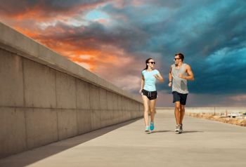 fitness, sport and people concept - happy couple in sports clothes and sunglasses running along pier over sunset sky on background. couple in sports clothes running along pier