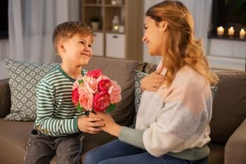mother’s day, holidays and family concept - happy little son giving flowers to his mother at home in evening. smiling little son gives flowers to mother at home