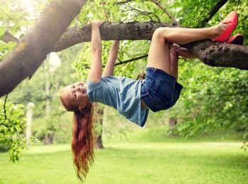 friendship, childhood, leisure and people concept - happy smiling little redhead girl hanging upside down on tree in summer park. happy little girl hanging on tree in summer park
