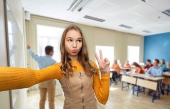 school, education and people concept - funny teenage student girl taking selfie and making faces over classroom background. teenage student girl taking selfie at school