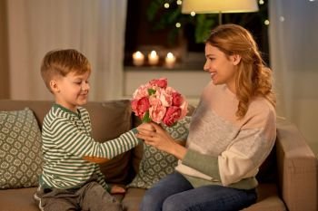 mother’s day, holidays and family concept - happy little son giving flowers to his smiling mother at home in evening. smiling little son gives flowers to mother at home