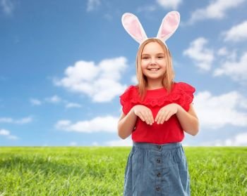 easter, holidays and childhood concept - happy girl wearing bunny ears headband over blue sky and grass background. happy girl wearing easter bunny ears headband