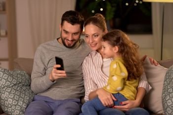 people, family and technology concept - happy mother, father and little daughter with smartphone at home at night. happy family with smartphone at home at night