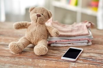babyhood, technology and clothing concept - baby clothes, teddy bear toy and smartphone on wooden table at home. baby clothes, teddy bear toy and smartphone