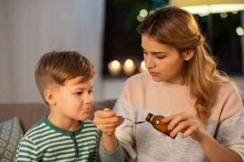 family, health and people concept - sad mother pouring antipyretic medication or cough syrup to spoon for ill little son at home. mother giving medication or cough syrup to ill son