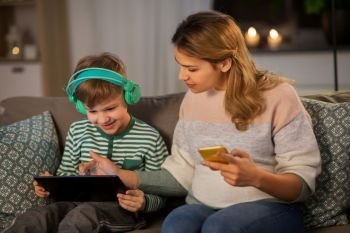 family, technology and people concept - happy mother and little son sitting on sofa with tablet pc computer, headphones and smartphone at home. mother and son using gadgets at home
